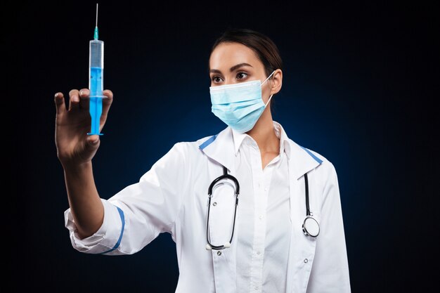 Young serious lady in medical mask holding and looking at syringe