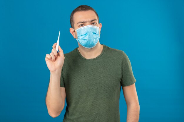 Young serious looking man wearing face medical mask holding a digital thermometer in raising hand isolated on blue