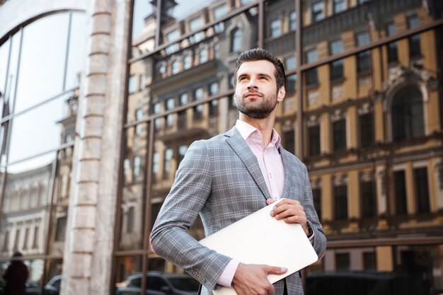 Young serious man in jacket holding laptop computer while standing