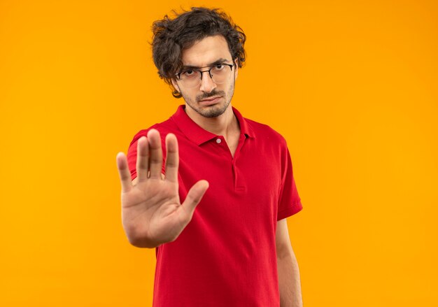 Молодой серьезный человек в красной рубашке с оптическими очками жестами останавливает знак рукой, изолирован на оранжевой стене