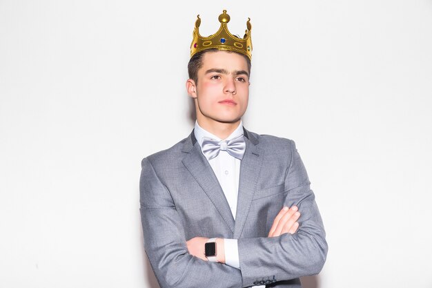 Young serious man in suit and tie, holding golden crown over his head, on white wall