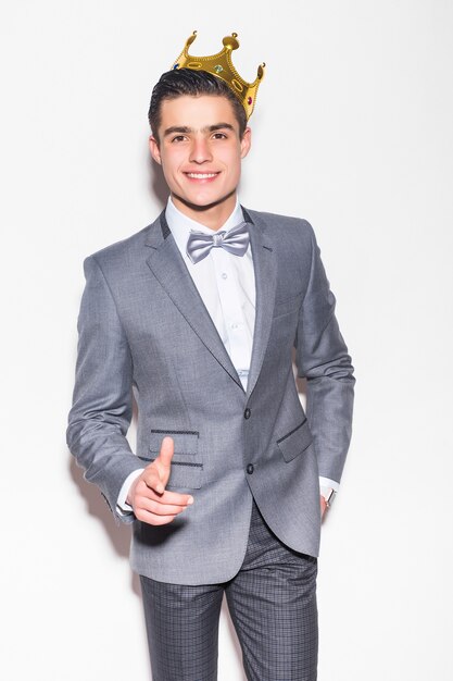 Young serious man in suit and tie, holding golden crown over his head, on white wall