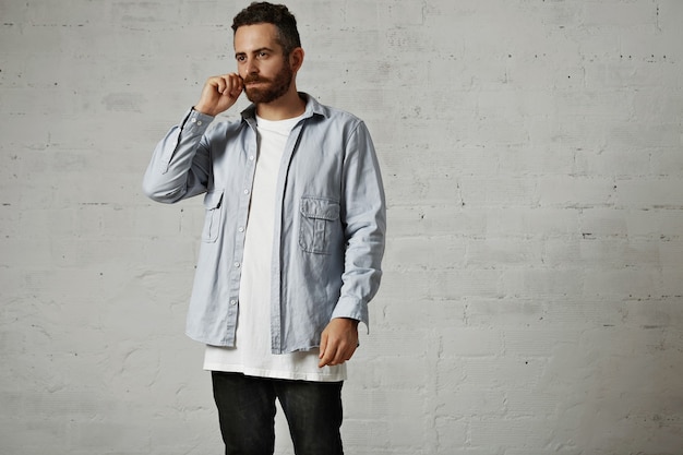 Young serious model wearing a white t-shirt, denim button-up shirt and black jeans adjusts his moustache isolated on white