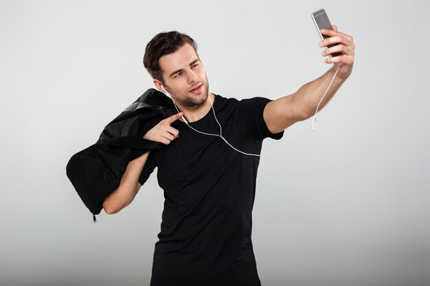 Молодой серьезный спортсмен делает селфи с сумкой по мобильному телефону