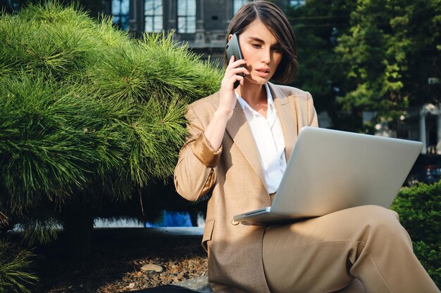 Young serious stylish businesswoman talking on cellphone and working on laptop outdoor