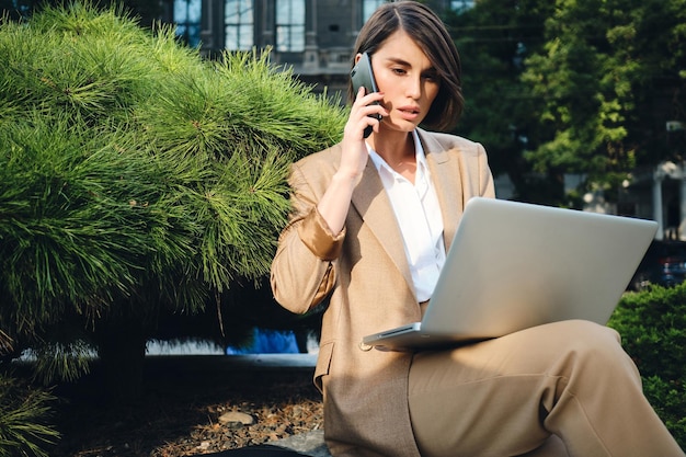 Молодая серьезная стильная деловая женщина разговаривает по мобильному телефону и работает на ноутбуке на открытом воздухе