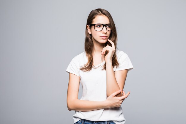 La giovane signora di pensiero serio in vetri in maglietta bianca e blue jeans rimane davanti al fondo bianco dello studio