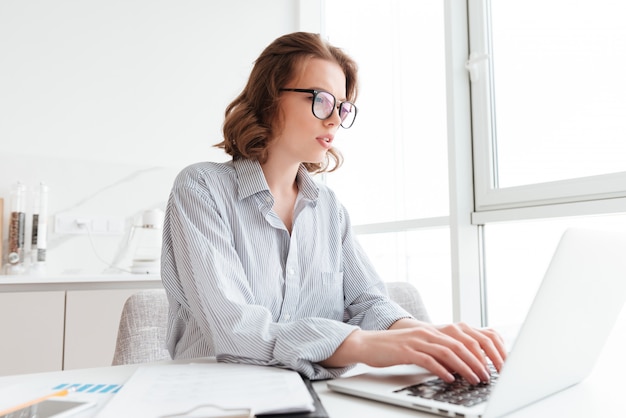 Young serious woman in striped shirt typing email to her boss while sitting at workplace in light apartment