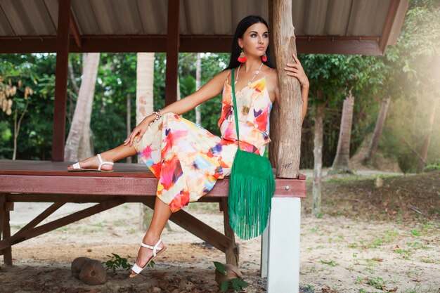 Young sexy beautiful woman in colorful dress, summer hippie style, tropical vacation, tanned legs, sandals, green handbag with fringe, accessories, smiling, happy