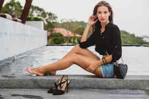 Free photo young sexy beautiful woman sitting at pool barefoot, long skinny tanned legs, high heeled shoes, black shirt, denim shorts, purse, relaxing, summer style, vacation, close-up details