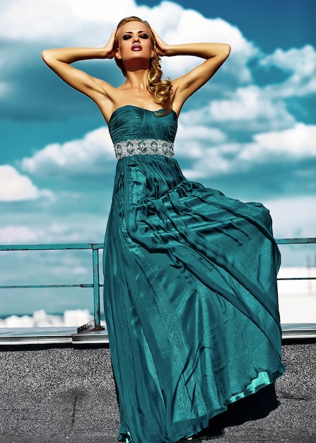 Young sexy blond woman model  in evening dress posing on blue sky background