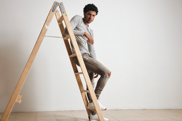 Young sexy latin guy wears blank grey henley longsleeve and jeans sits on wooden ladder , isolated on white