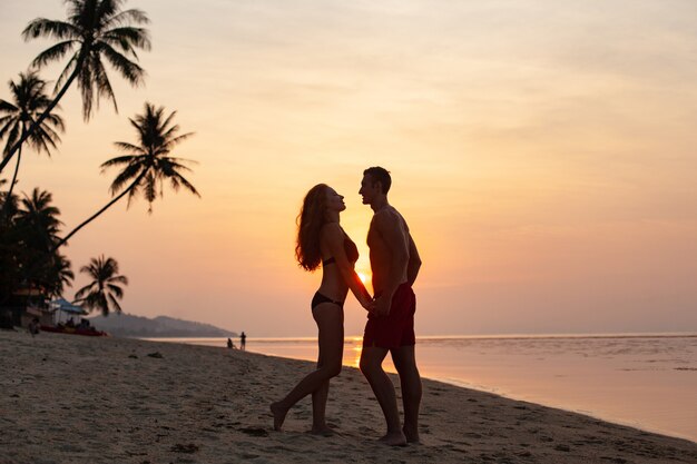 Молодая сексуальная романтическая пара в любви на закате счастлива на летнем пляже вместе весело в купальных костюмах
