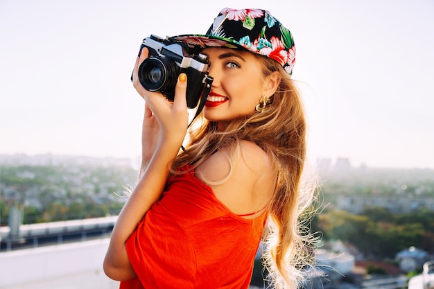若いセクシーな官能的なブロンドの女性はレトロなビンテージヒップスターカメラで写真を撮って、笑って楽しんで、盗品の花の明るい帽子をかぶっています。