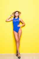 Free photo young sexy slim tanned woman in blue swimsuit posing against yellow wall. swimwear or bikini model