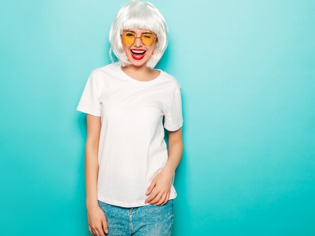 Young sexy smiling hipster girl in white wig and red lips.Beautiful trendy woman in summer clothes.Carefree model posing near blue wall in studio summer  going crazy in round sunglasses