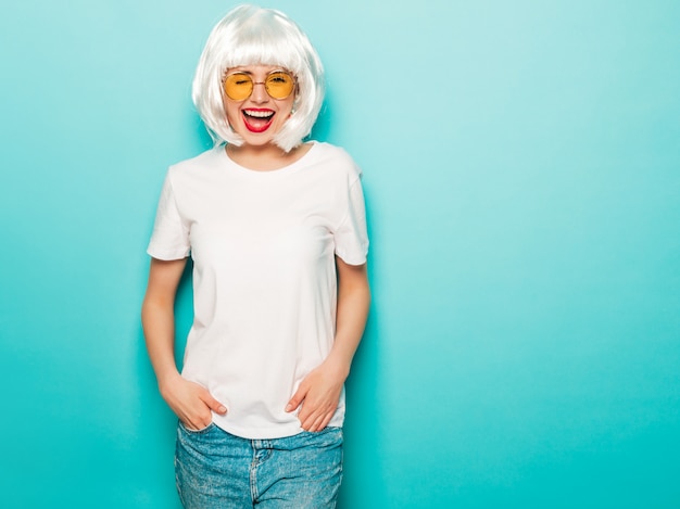 Young sexy smiling hipster girl in white wig and red lips.Beautiful trendy woman in summer clothes.Carefree model posing near blue wall in studio summer  going crazy