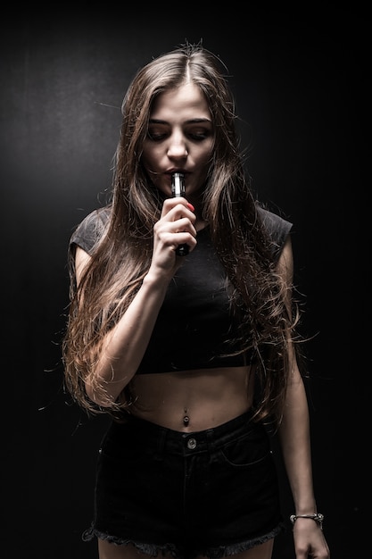 若いセクシーな女性はvaping喫煙電子タバコです。黒い壁に蒸気の雲