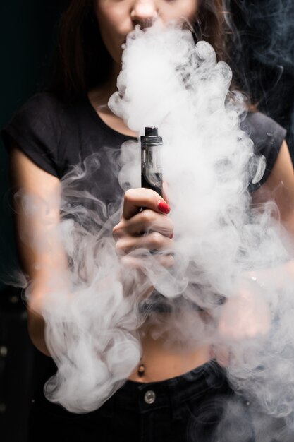 Молодая сексуальная женщина vaping курить электронную сигарету. облако пара на черной стене