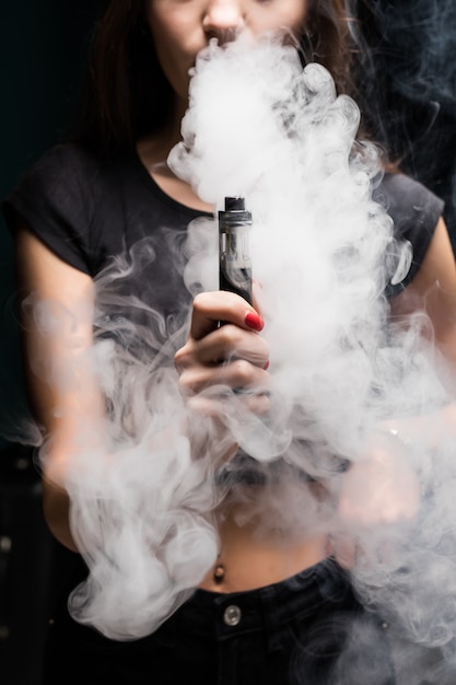 젊은 섹시 한 여자는 담배 전자 담배를 vaping입니다. 검은 벽에 증기의 구름