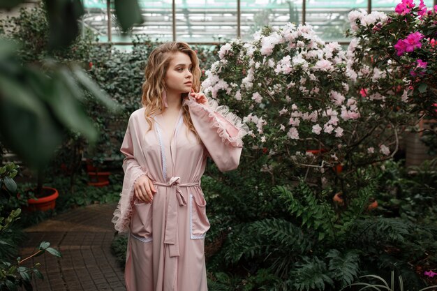 花がいっぱいの朝の庭に立っているピンクのバスローブで若いセクシーな女性