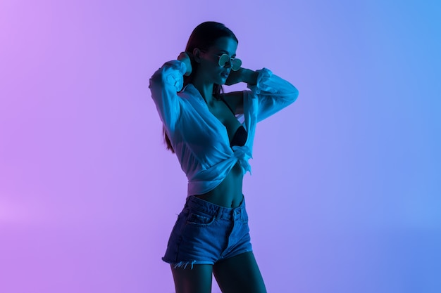 Young sexy woman in short and shirt, sunglasses standing isolated on pink light