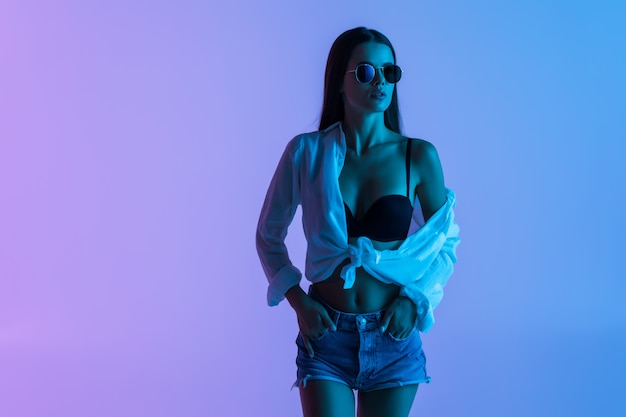 若いセクシーな女性の短いシャツとピンクの光に分離されて立っているサングラス