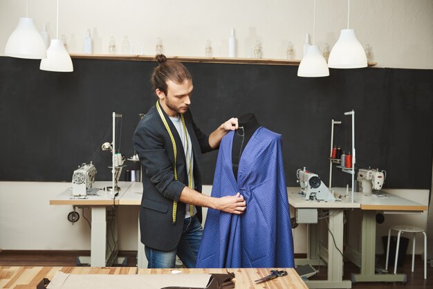 Young shaved good-looking caucasian male fashion designer in stylish outfit working on new blue dress for spring collection in his workshop. Artist creating beautiful clothes in his workshop