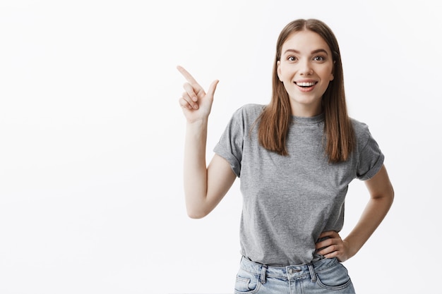 Young sheerful beautiful dark-haired young woman with medium hair lenghts in grey t-shirt and jeans smiling brightfully,  with sutisfied and happy expression, pointing at white wall.