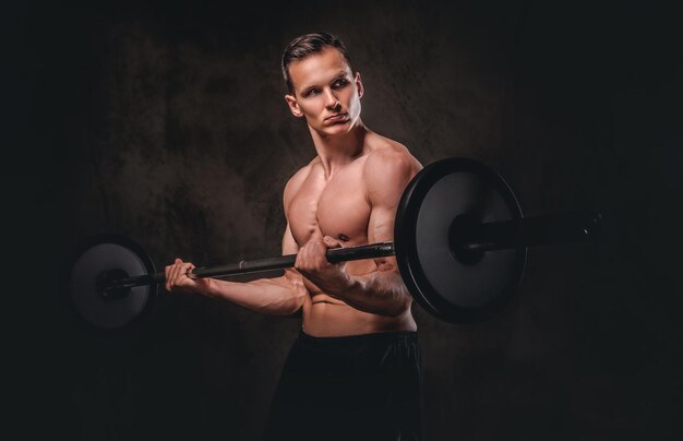 Young shirtless bodybuilder holding a barbell and doing exercise on biceps. Isolated on a dark background.