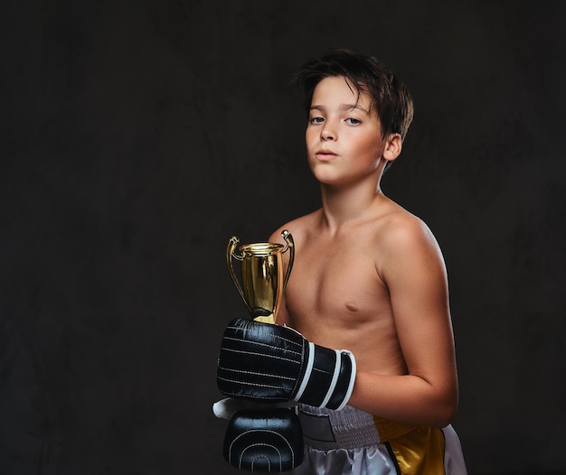 Young shirtless boxer champion wearing gloves holds a winner's cup. Isolated on a dark background.