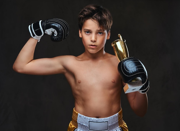 手袋をはめた若い上半身裸のボクサーチャンピオンは、筋肉を示す勝者のカップを保持しています。暗い背景に分離されています。