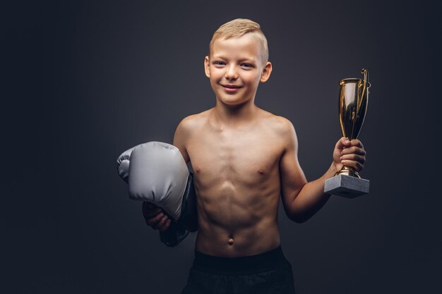 Молодой мальчик без рубашки держит боксерские перчатки и кубок победителя. Изолированные на темном фоне.