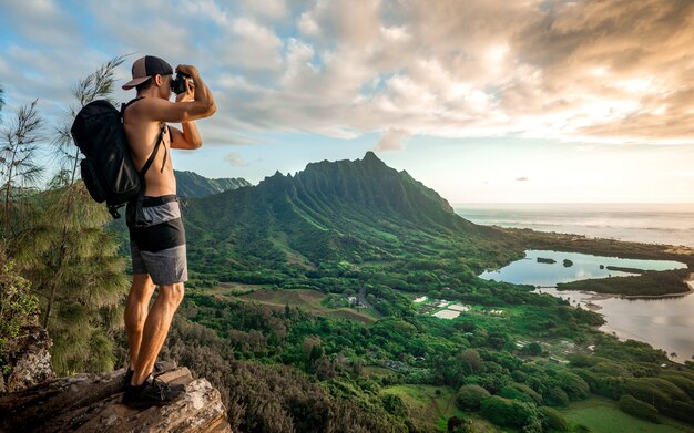 山の上に立って曇り空の下で写真を撮るバックパックを持つ若い上半身裸の男性