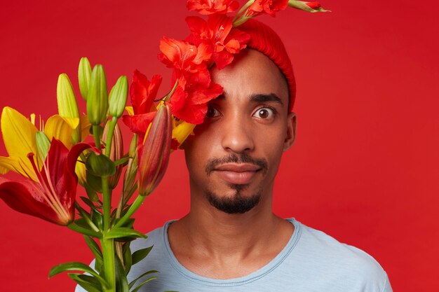 Young shocked attractive guy in red hat and blue t-shirt, holds a bouquet in his hands and covered part of face with flowers, looks at the camera with wide open eyes, stands over red backgroud