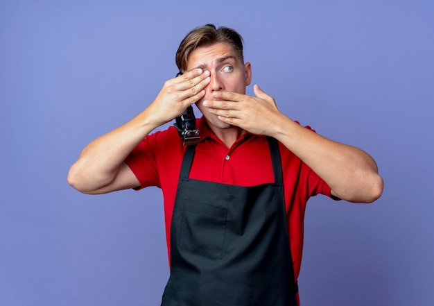 Foto gratuita il giovane barbiere maschio biondo scioccato in uniforme chiude l'occhio con la mano mette la mano sulla bocca che tiene il tagliatore di capelli e guardando il lato isolato sullo spazio viola con lo spazio della copia