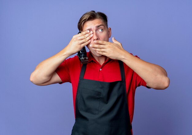 Young shocked blonde male barber in uniform closes eye with hand puts hand on mouth holding hair clipper and looking at side isolated on violet space with copy space