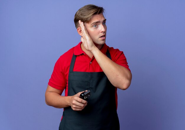 Young shocked blonde male barber in uniform hides face with hand looking at side holding hair clipper isolated on violet space with copy space