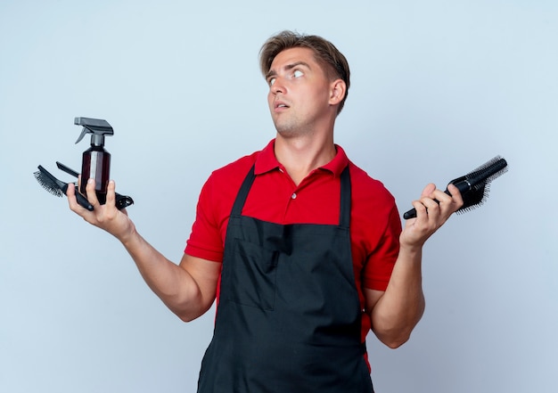 Young shocked blonde male barber in uniform holds barber tools looking at side isolated on white space with copy space