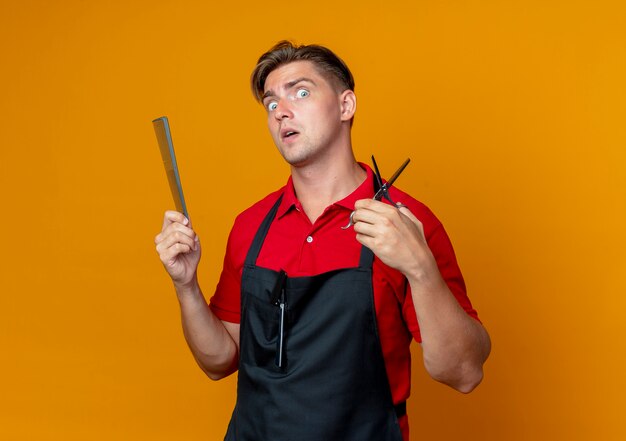 Young shocked blonde male barber in uniform holds comb and scissors isolated on orange space with copy space