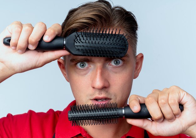 young shocked blonde male barber in uniform holds hair combs on forehead and chin looking at camera