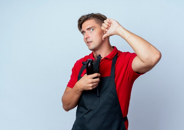 Young shocked blonde male barber in uniform thumbs down holding barber tools isolated on white space with copy space