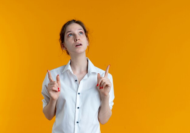 Young shocked blonde russian girl looks and points up with two hands isolated on orange space with copy space