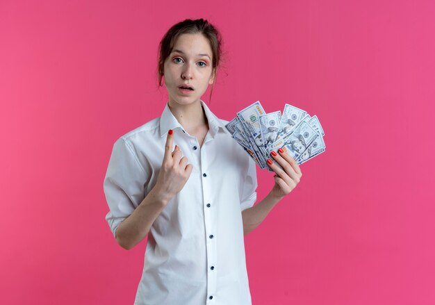 Young shocked blonde russian girl points up holding money isolated on pink space with copy space