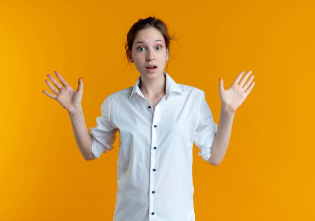 Young shocked blonde russian girl stands with raised hands isolated on orange space with copy space