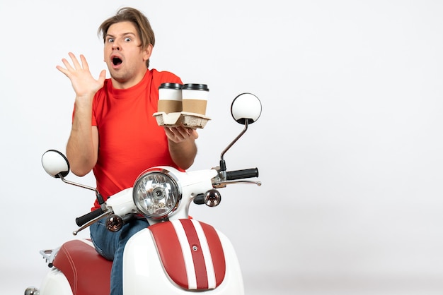 Ragazzo giovane corriere scioccato in uniforme rossa che si siede sul motorino che tiene gli ordini sulla parete gialla