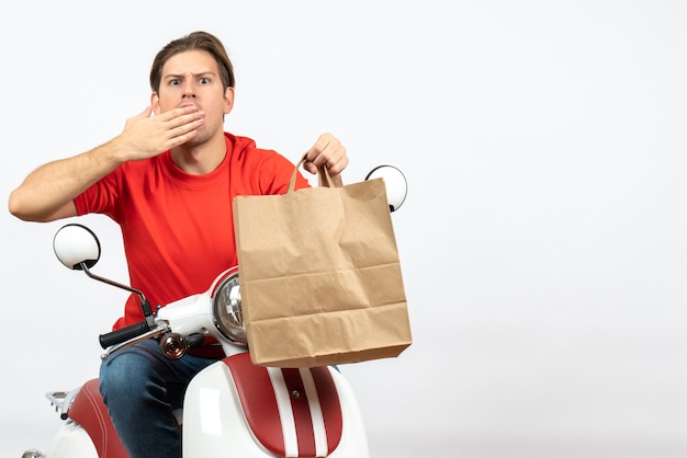 Young shocked courier guy in red uniform sitting on scooter holding paper bag and putting hand on his mouth on white wall