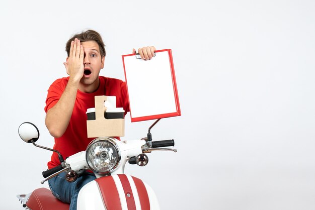 Young shocked courier man in red uniform sitting on scooter holding orders and documents closing his eye on yellow wall