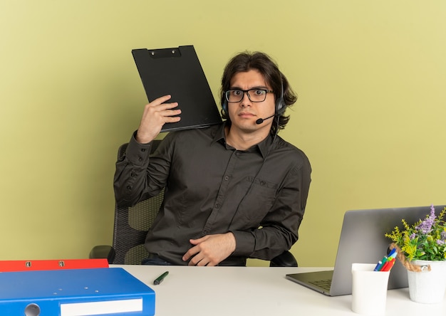 Foto gratuita giovane scioccato ufficio lavoratore uomo con le cuffie si siede alla scrivania tiene appunti guardando la fotocamera