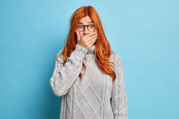 若いショックを受けた赤毛の若い女性が口を覆い、恥ずかしいニュースが透明なメガネの灰色のニットセーターを着ているのを聞いて愚かな視線を向けます。